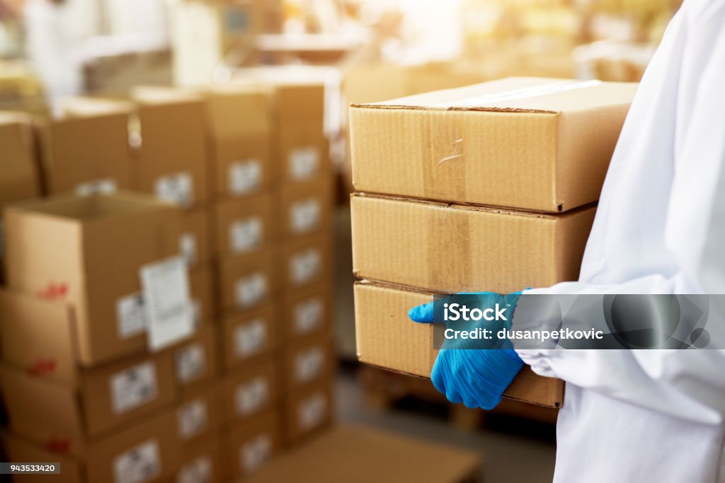 Close up view of a dedicated worker carrying a stack of duck taped brown boxes in factory storage room while wearing sterile cloths and rubber gloves. Healthcare And Medicine Stock Photo