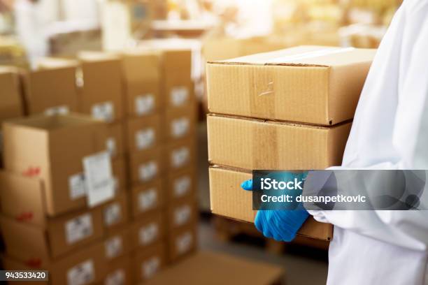 Close Up View Of A Dedicated Worker Carrying A Stack Of Duck Taped Brown Boxes In Factory Storage Room While Wearing Sterile Cloths And Rubber Gloves Stock Photo - Download Image Now