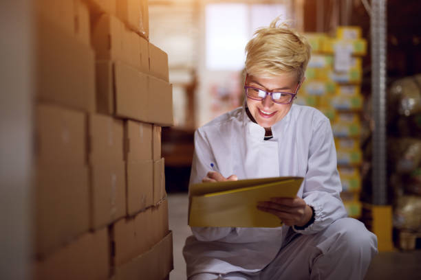 istock Young cheerful female worker in facility storage area is checking box stacks and filling a paperwork in a yellow folder. 943539042