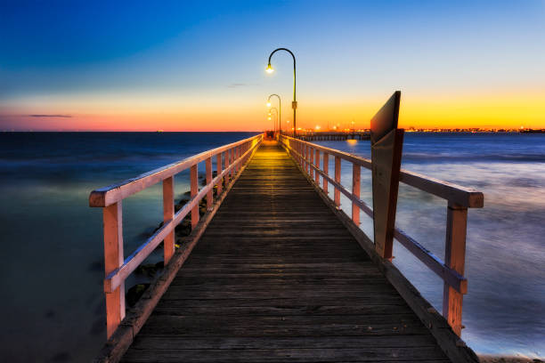 Me Port Jetty Centre set Lagoon pier made of timber in Port Melbourne at sunset. Empty abandoned sandy beach on Port Phillip bay of Melbourne city facing orange sky of setting sun. port melbourne melbourne stock pictures, royalty-free photos & images