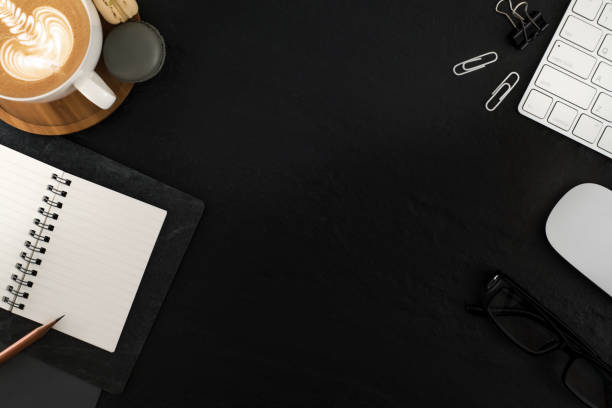 istock Flat lay, top view office table desk. Workspace with blank note book, keyboard, macaroon, office supplies, eyeglass and coffee cup on black stone background. 943588410
