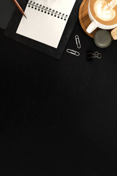 istock Flat lay, top view office table desk. Workspace with blank note book, keyboard, macaroon, office supplies, eyeglass and coffee cup on black stone background. 943588412