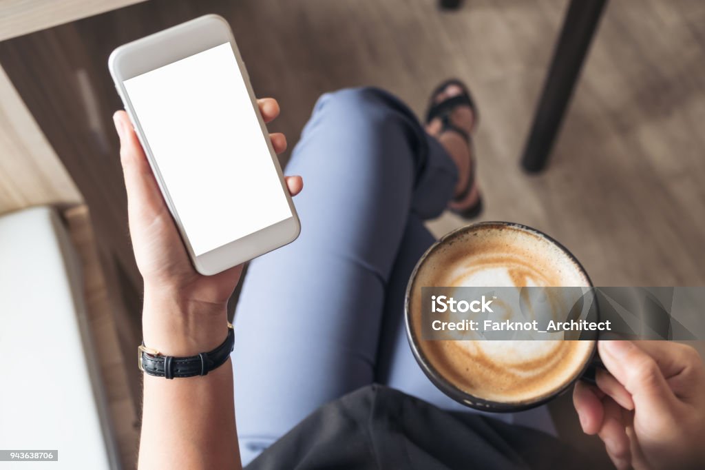 Top view mockup image of a woman's hand holding white mobile phone with blank desktop screen while drinking coffee in cafe Coffee - Drink Stock Photo