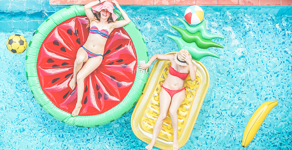 Happy girls floating with tropical fruit lilos inside swimming pool - Young women friends relaxing in summer vacation at resort hotel - Travel, chilling, holidays, youth concept - Focus on heads hats