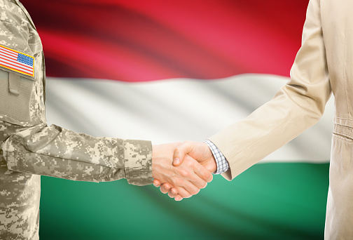 American soldier in uniform and civil man in suit shaking hands with adequate national flag on background - Hungary