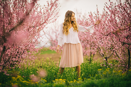 Young beautiful hipster woman near sakura blooming tree. Pink flowers, spring and youth concept. Stylish girl smiling. Blooming garden with cherry-tree