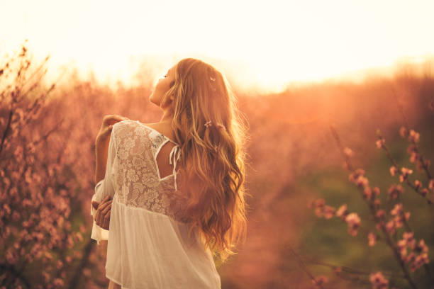 Portrait of beautiful young woman with healthy skin who enjoying life and standing among cherry branches with flower blossom at sunset. She is very happy and cheerful