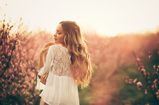 Portrait of beautiful young woman with healthy skin who enjoying life and standing among cherry branches with flower blossom at sunset. She is very happy and cheerful
