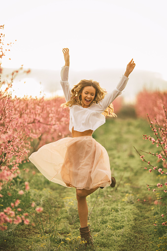 Very happy carefree woman standing among cherry threes with flower blossoms at spring. She is jumping, dancing, enjoying life