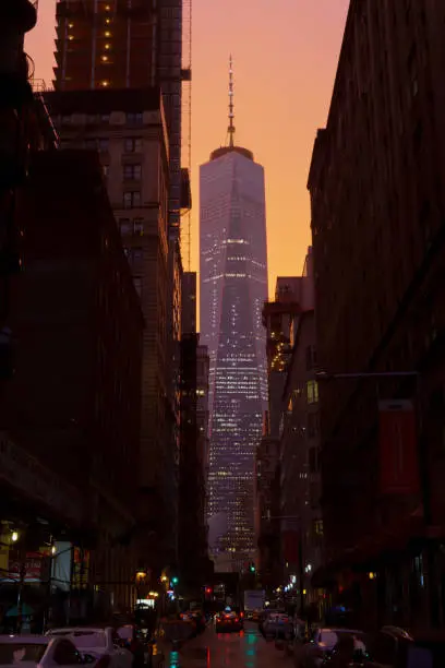 A summer rainstorm leaves One World Trade Center illuminated in purple with a peach-colored sky. Scaffolding, cars, a taxicab, and stores can be seen surrounding it on Fulton Street below.