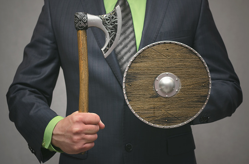 Businessman holds in hand a toy viking axe weapon and shield. Life or finance insurance agent. Money deposite safety agent.
