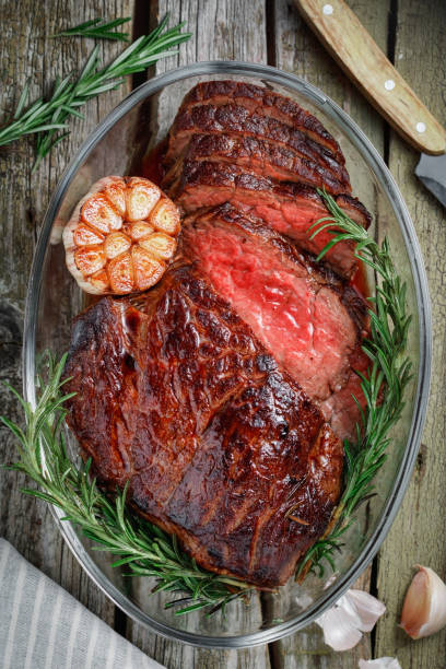 Top view of a baked beef steak medium rare cut into pieces in a glass bowl with baked garlic and rosemary. The concept of a festive table and dishes.
