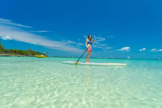 A woman on a standup paddlboard  in shallow clear waters near a beach on Eleuthera Island, yachts anchored in background.