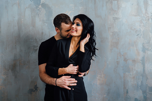 Young stylish beautiful gorgeous couple, against the gray wall loft in the studio or at home. Valentine's Day. black fashionable clothes. Exquisite clothes, dark dress. Kisses. Hugs from behind.