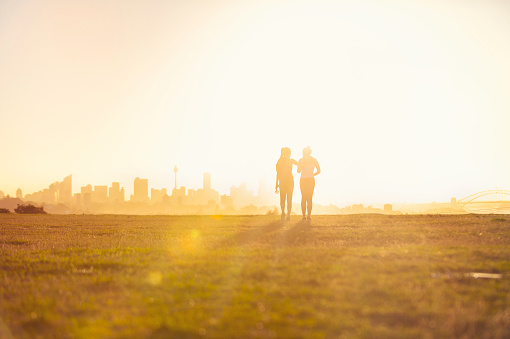 Silhouette of 2 women walking in the park. They are exercising an about to go running at sunset or sunrise with the city of Sydney in the background. One woman has her arm around the other. Could be a lesbian couple. Copy space