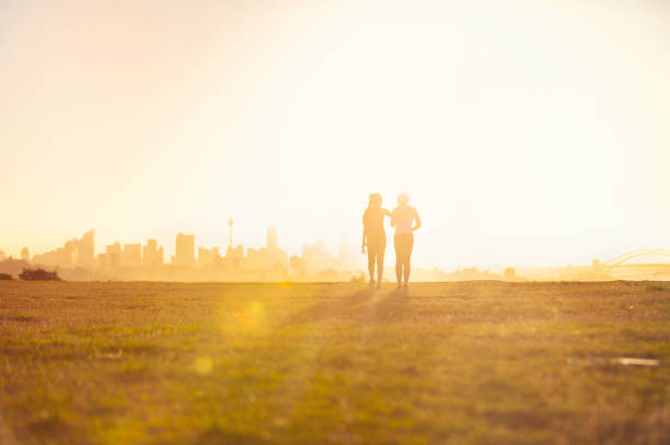 Silhouette of 2 women walking in the park. Silhouette of 2 women walking in the park. They are exercising an about to go running at sunset or sunrise with the city of Sydney in the background. One woman has her arm around the other. Could be a lesbian couple. Copy space lesbian couple embracing stock pictures, royalty-free photos & images