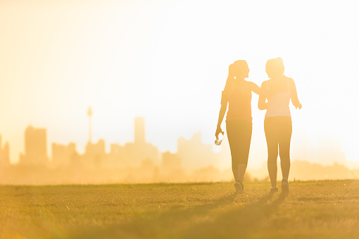 Silhouette of 2 women walking in the park. They are exercising an about to go running at sunset or sunrise with the city of Sydney in the background. One woman has her arm around the other. Could be a lesbian couple. Copy space
