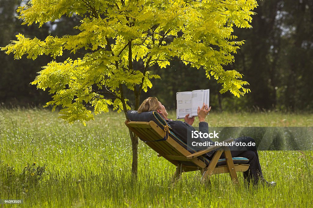 Mature woman lying outdoors in deck chair reading book Mature woman lying on wooden deck chair in grass under small golden acacia tree reading book. Adult Stock Photo