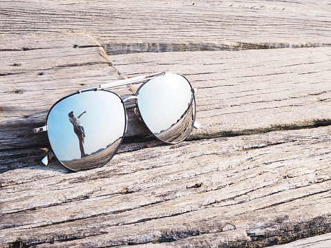Reflection of woman hand pointing upward to blue sky in fashionable mirror sunglasses on vintage wood background. Happy summer vacation holiday concept.