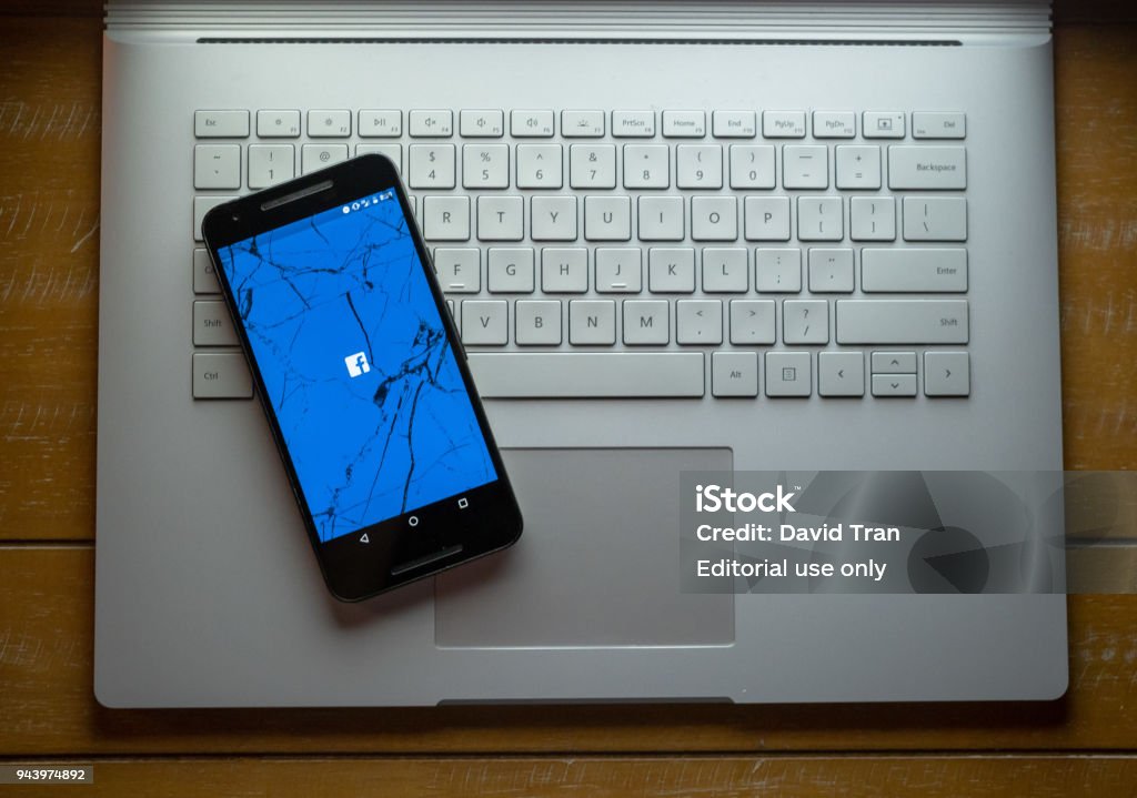 A cracked phone with the Facebook app loading sitting on a laptop APRIL 8, 2018: A cracked phone with the Facebook app loading sitting on a laptop. The social media giant has been in hot water for privacy troubles. Cambridge Analytica Stock Photo