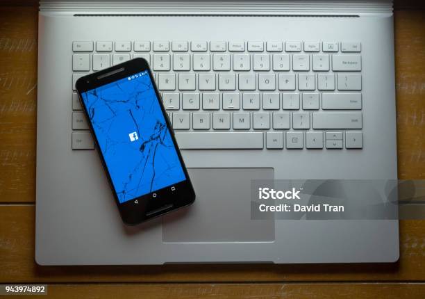 A Cracked Phone With The Facebook App Loading Sitting On A Laptop Stock Photo - Download Image Now