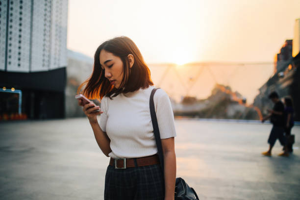 Vintage colored portrait of a young Asian woman, relaxing and texting on the cellphone at the busy Siam station of the Skytrain, next to popular shopping malls and restaurants. She is wearing casual clothing, drinking a juice to refresh.