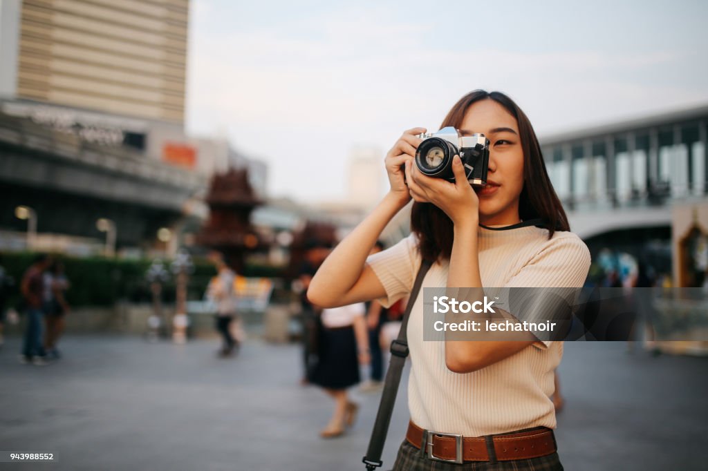 Young Asian woman traveler in Bangkok downtown district, holding a vintage film camera Vintage colored portrait of a young Asian woman traveler, relaxing at the busy Siam station of the Skytrain, next to popular shopping malls and restaurants. She is wearing casual clothing, taking photos with her beautiful analog film camera with a fixed, large aperture, prime lens. Photographer Stock Photo