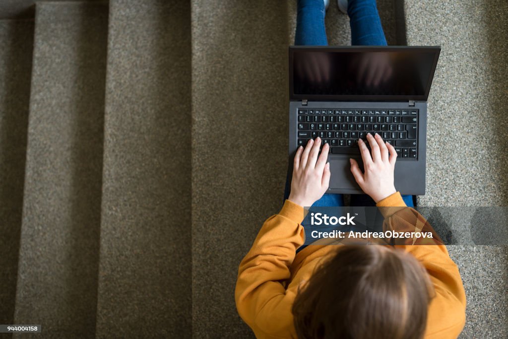 若い女子の学生の学校で階段に座っている彼女のラップトップ上のエッセイを書きます。教育コンセプトです。上からの眺め。 - ノートパソコンのロイヤリティフリーストックフォト