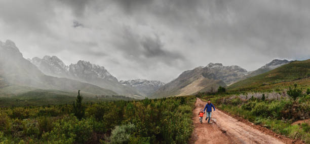 Father and Son Running Together between snow capped mountains A panoramic photo of a father and 2-3 year old son with a bright orange coloured down jacket holding hands running happily on a dirt road in Jonkershoek Stellenbosch with the mountains covered in snow and a dramatic sky fynbos photos stock pictures, royalty-free photos & images