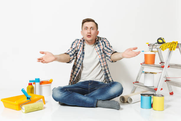 istock Confused shocked annoyed tired man spreading hands, sitting on floor with instruments for renovation apartment isolated on white background. Wallpaper, gluing accessories, tools. Repair home concept. 944038592