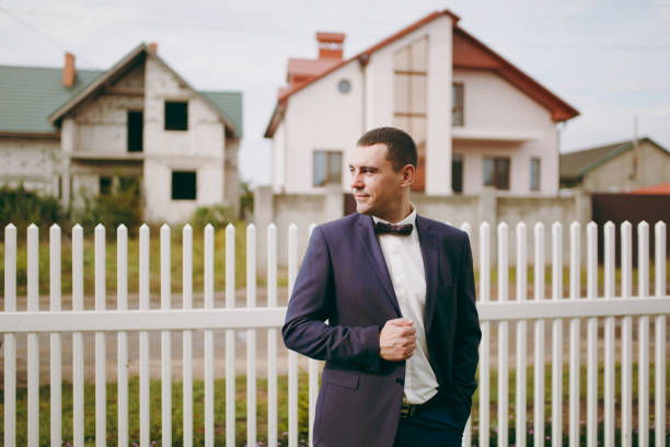 Morning preparation of the newlyweds for the wedding ceremony. The stylish smiling groom in blue formal suit, white shirt and bow tie standing on the background of fence and houses. Wedding wear Morning preparation of the newlyweds for the wedding ceremony. The stylish smiling groom in blue formal suit, white shirt and bow tie standing on the background of fence and houses. Wedding wear niñas stock pictures, royalty-free photos & images