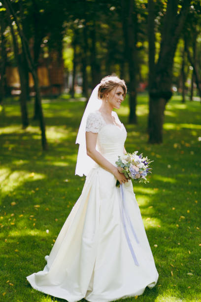 Beautiful wedding photosession. Elegant young bride in white dress and veil with beautiful hairdress with bouquet of flowers and ribbons near trees on wedding walk in the big green park on sunny day Beautiful wedding photosession. Elegant young bride in white dress and veil with beautiful hairdress with bouquet of flowers and ribbons near trees on wedding walk in the big green park on sunny day. niñas stock pictures, royalty-free photos & images