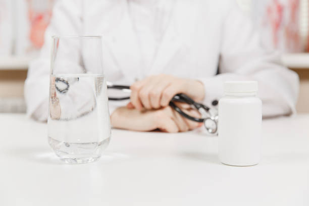 Doctor at desk with bottle white pills, glass of water in office in hospital. Woman holds in hands stethoscope in consulting room. Medicine concept. Copy space, with place for text. Advertising area. Doctor at desk with bottle white pills, glass of water in office in hospital. Woman holds in hands stethoscope in consulting room. Medicine concept. Copy space, with place for text. Advertising area niñas stock pictures, royalty-free photos & images