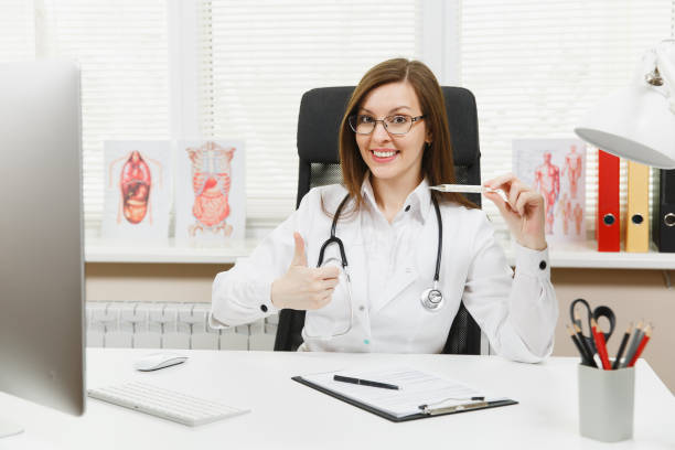 Female doctor sitting at desk with medical documents, clinical thermometer with normal temperature in light office in hospital. Woman in medical gown, stethoscope in consulting room. Medicine concept. Female doctor sitting at desk with medical documents, clinical thermometer with normal temperature in light office in hospital. Woman in medical gown, stethoscope in consulting room. Medicine concept niñas stock pictures, royalty-free photos & images