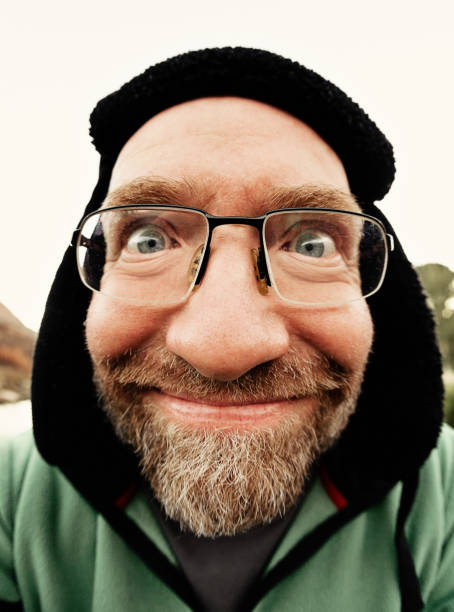 A mature man wearing spectacles  and a winter hat with ear flaps grins as he takes an outdoor selfie.