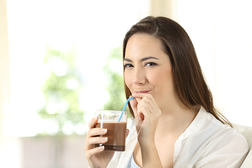Woman drinking cocoa shake with a straw sitting on a couch in the living room at home