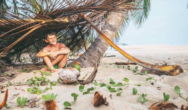 istock Boy sits in selfmade hut on the tropical beach and plays in Robinzone 944097980