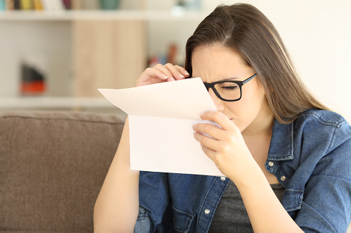 Woman having eyesight problems to read a letter sitting on a couch in the living room at home