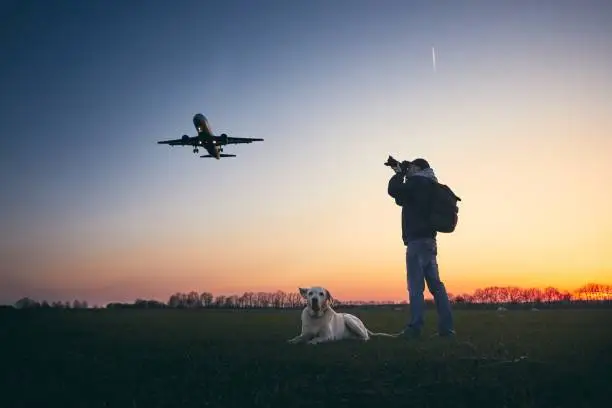 Young man with dog is photographing near airport. Airplane landing aginst moody sky sky at golden sunset.