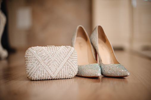 graceful shoes of the bride, studded with shiny pebbles, stand on the beige floor next to a small white clutch decorated with beads