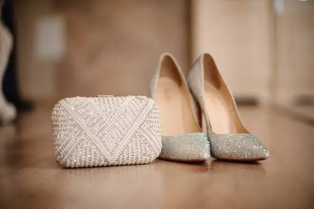 Photo of shoes of the bride with shiny pebbles stand next to a white clutch