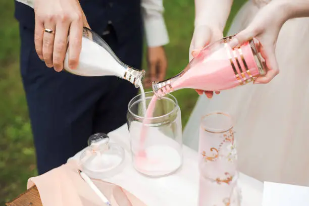 Closeup view of bride and groom during wedding sand ceremony. Pouring decorative white and pink sand into vase as symbol of unity. Horizontal color photography.