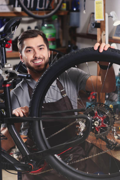istock handsome young man in apron fixing bicycle and smiling at camera in workshop 944222086