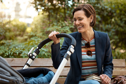Smiling mother looking in baby stroller. Mid adult woman with book and pram sitting in park. She is wearing blazer outdoors.