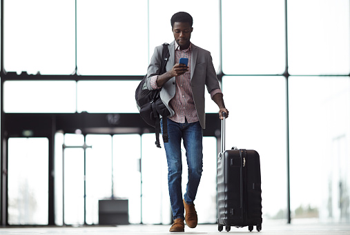 Young mobile traveler with smartphone, backpack and suitcase walking along airport and messaging or looking through online information