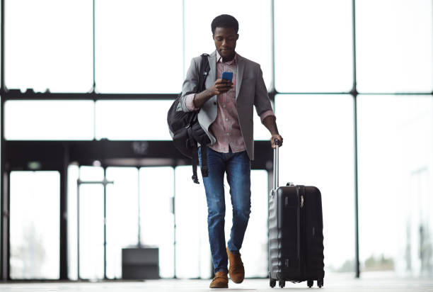 Texting on the move Young mobile traveler with smartphone, backpack and suitcase walking along airport and messaging or looking through online information businessman african descent on the phone business person stock pictures, royalty-free photos & images