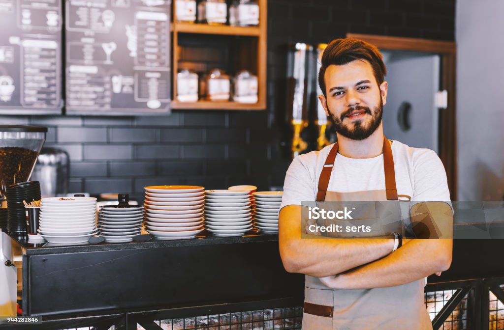 Good day start with coffee! Portrait of smiling young confident waiter handsone hipster barista man look at camera posing wearing apron standing behind the counter of a store with his arms folded in his coffee shop. xxxl size Barista Stock Photo