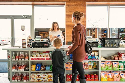 Mother with son shopping in a delicatessen store, being helped by smiling cashier.