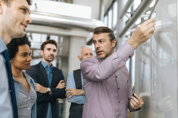 istock Mid adult entrepreneur talking to his colleagues while drawing business development on whiteboard. 944305056