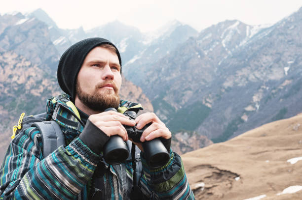 istock A hipster man with a beard in a hat, a jacket, and a backpack in the mountains holds binoculars, adventure, tourism, tracking 944310086